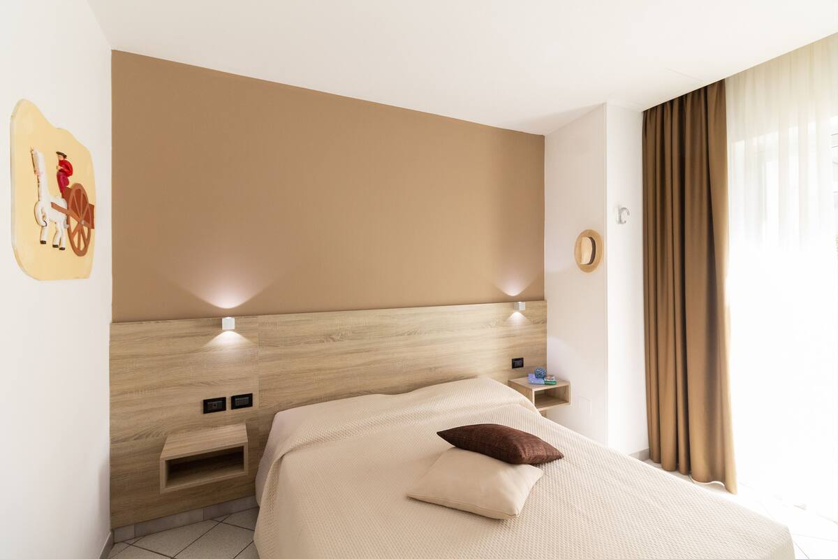 A modern hotel room with a wooden headboard, a bed with a beige bedspread and two pillows, a nightstand with a lamp, and a wall-mounted artwork.