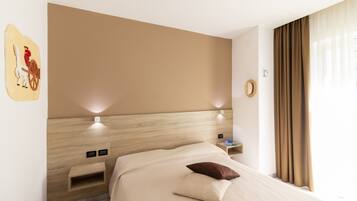 A modern hotel room with a wooden headboard, a bed with a beige bedspread and two pillows, a nightstand with a lamp, and a wall-mounted artwork.