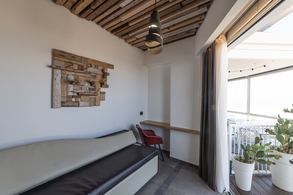 A balcony with a wooden ceiling, a cushioned bench, a red chair, a wooden wall art piece, and potted plants.