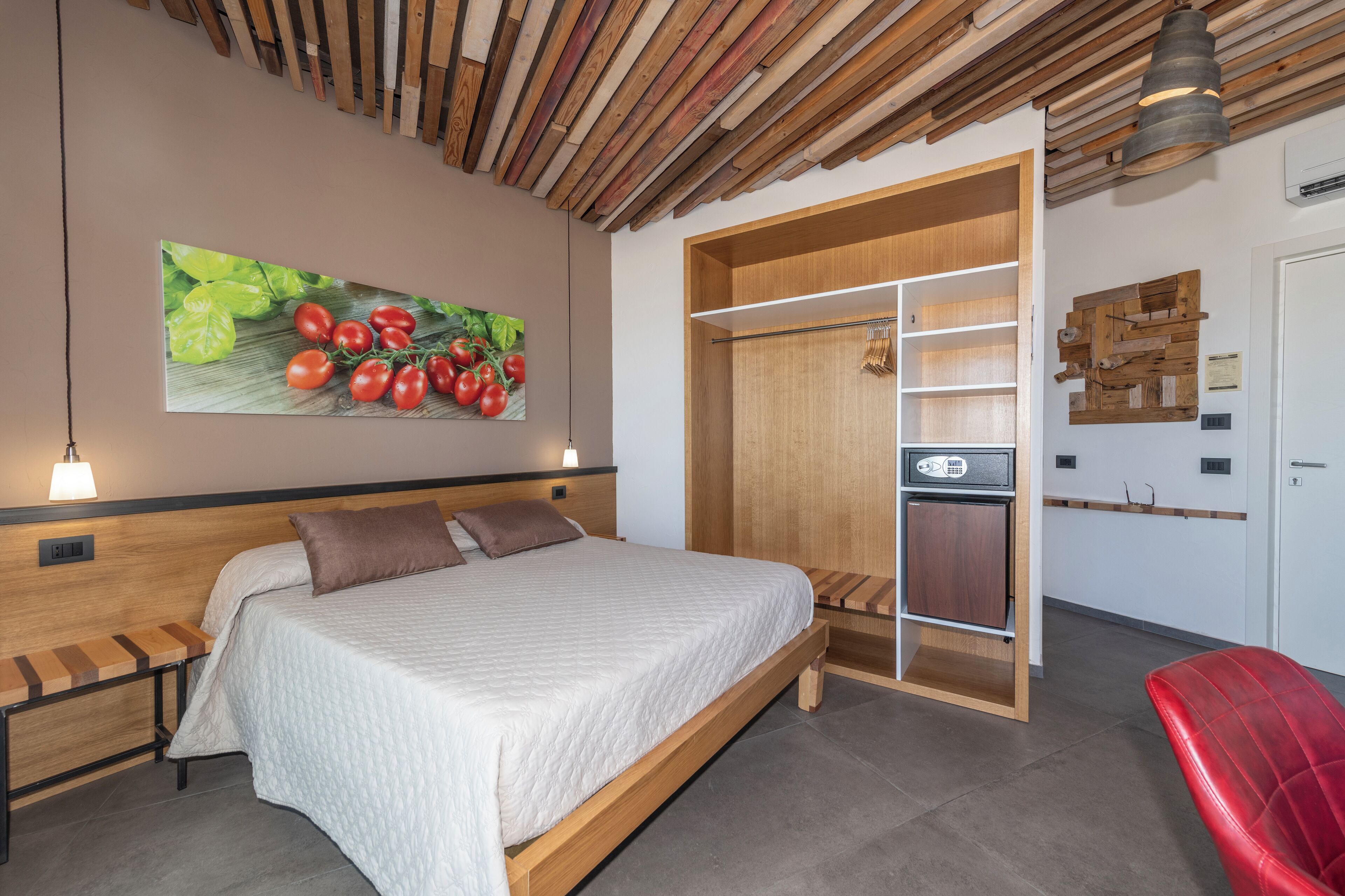 Un dormitorio moderno con cabecera de madera, una cama grande con colcha, un banco, una silla roja, un armario de madera y una estantería fijada a la pared.
