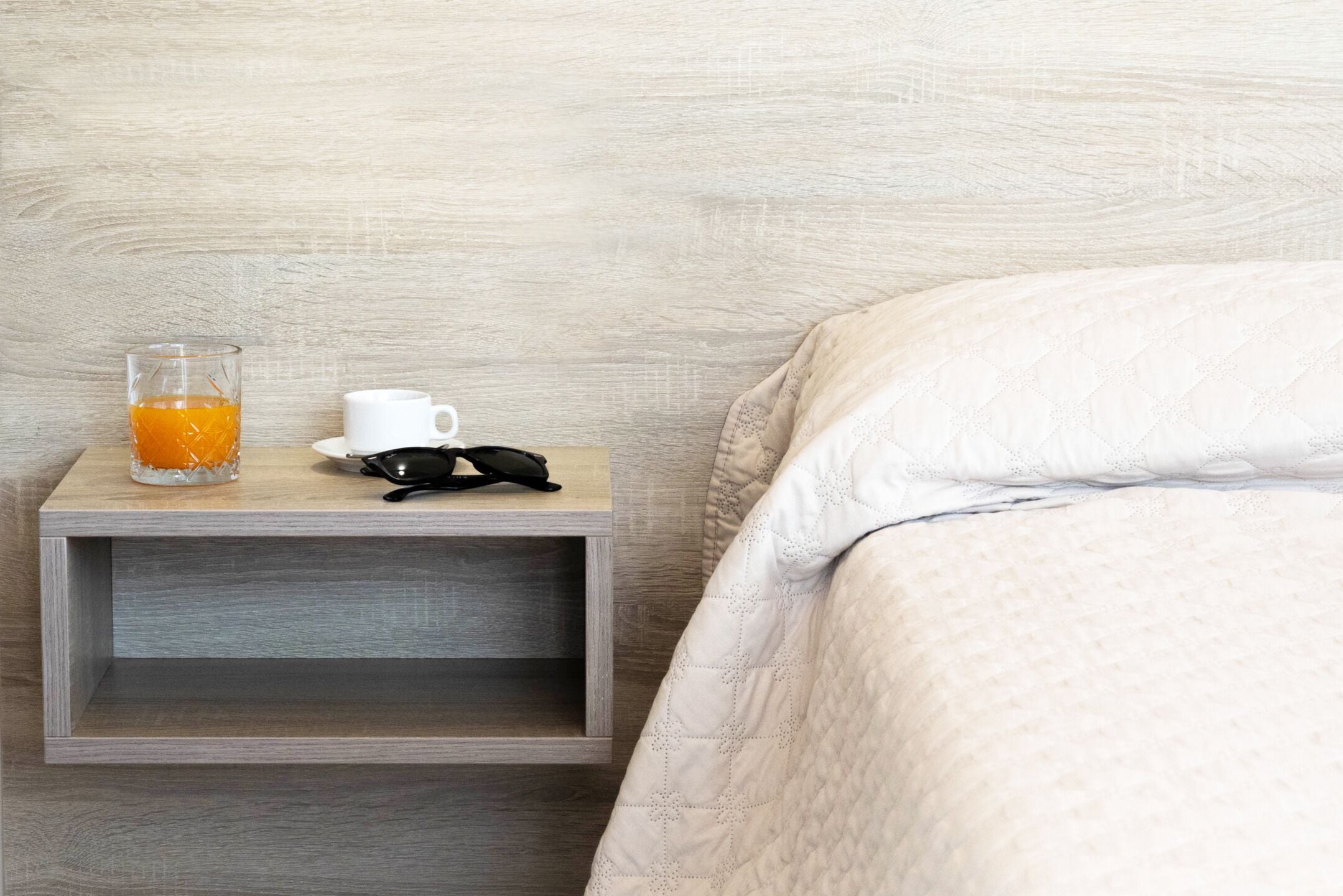 A wooden bedside table with a glass of orange juice, a white cup, and a pair of sunglasses.