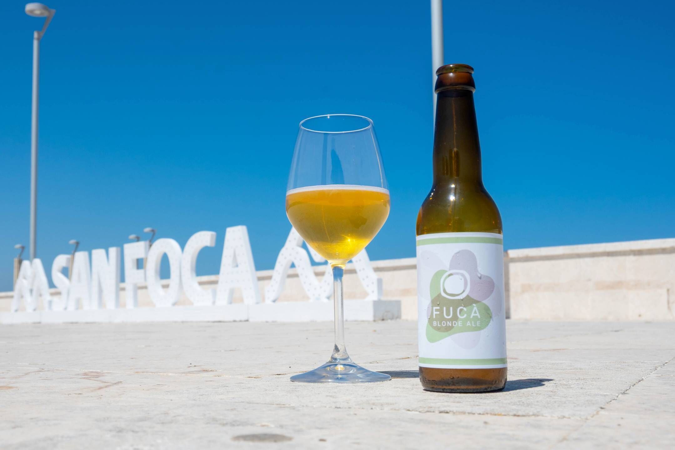 Une bouteille de Fuca Blonde Ale et un verre de bière posés sur une surface en béton, avec un ciel bleu clair en arrière-plan.