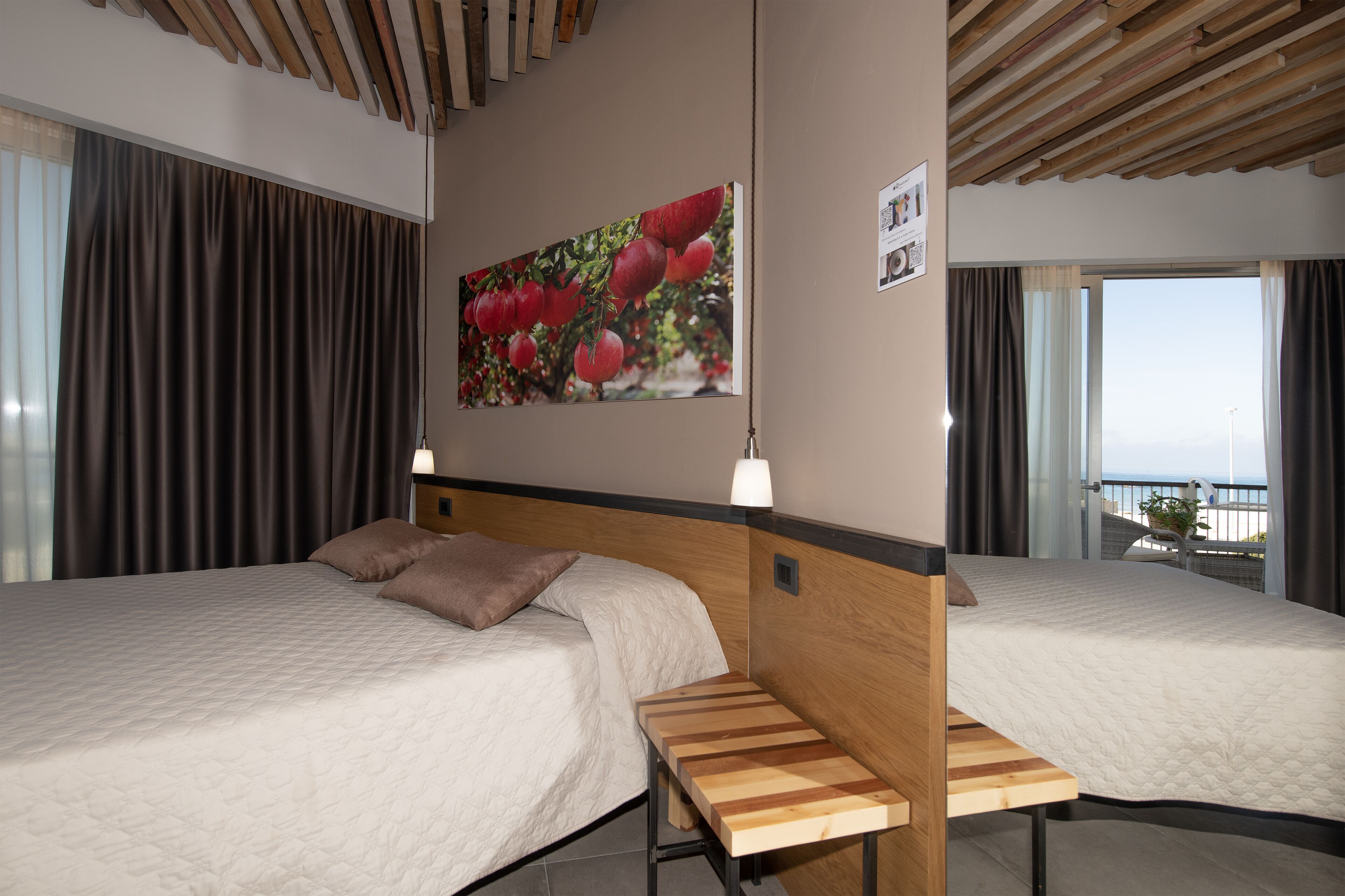 A hotel room with two beds, a wooden bench, a large fruit painting, and a balcony with a view.