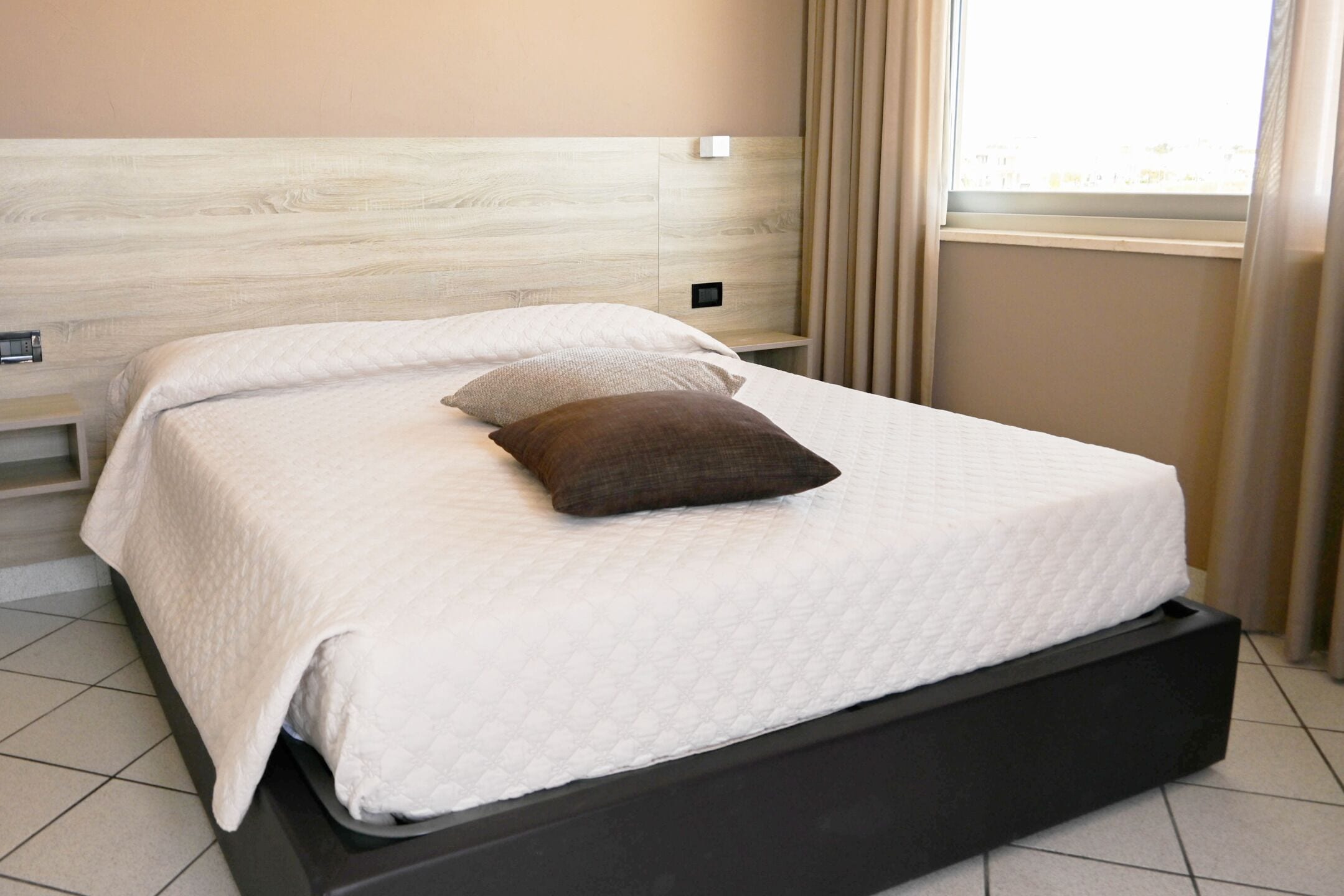 A neatly made bed with a white and brown pillow, a wooden headboard, and a window with curtains.