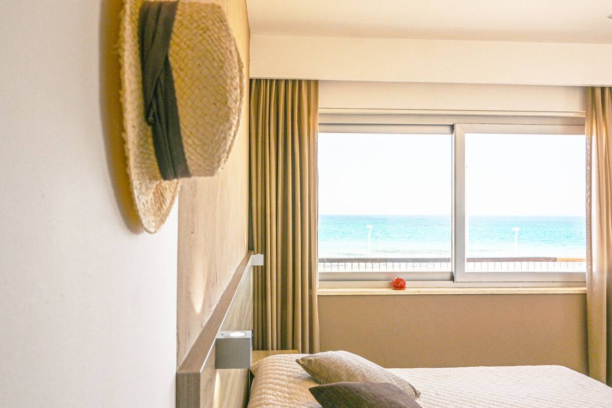 A hotel room with a bed, a window with a view of the sea, and a hat hanging on the wall.