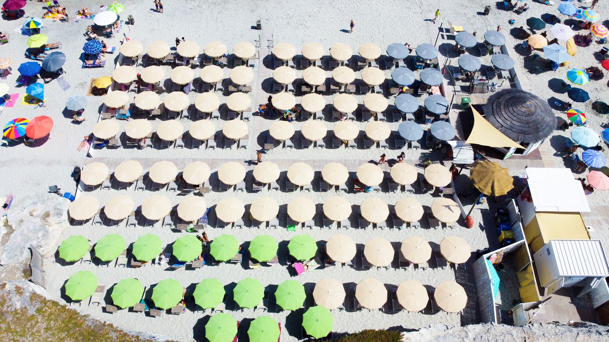 An aerial view of a beach with rows of sun loungers and umbrellas, some with people, and a few beach chairs.