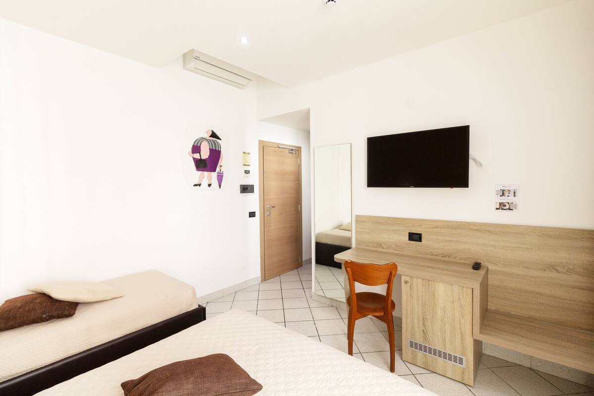 A hotel room with a bed, a desk, a chair, a TV mounted on the wall, and a door leading to another room.