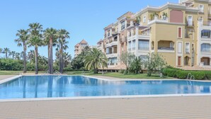 Une piscine dotée d’une terrasse carrelée, entourée de palmiers et bordée d’un immeuble à plusieurs étages avec des balcons.