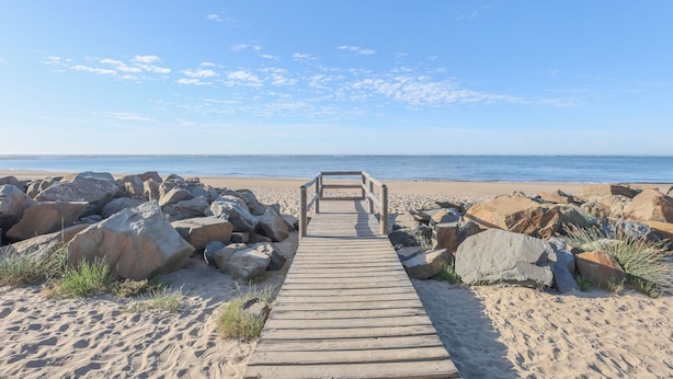 Un sentier en bois mène à une plage parsemée de gros rochers et où la végétation est rare.