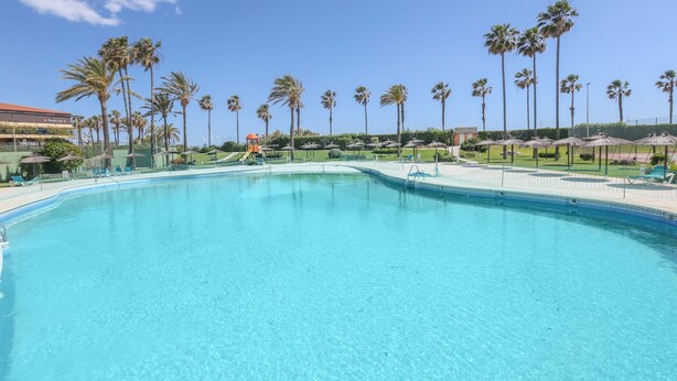 A clear blue swimming pool surrounded by palm trees and umbrellas, with a slide and lounge chairs visible. - Las Americas in Islantilla (Isla Cristina)