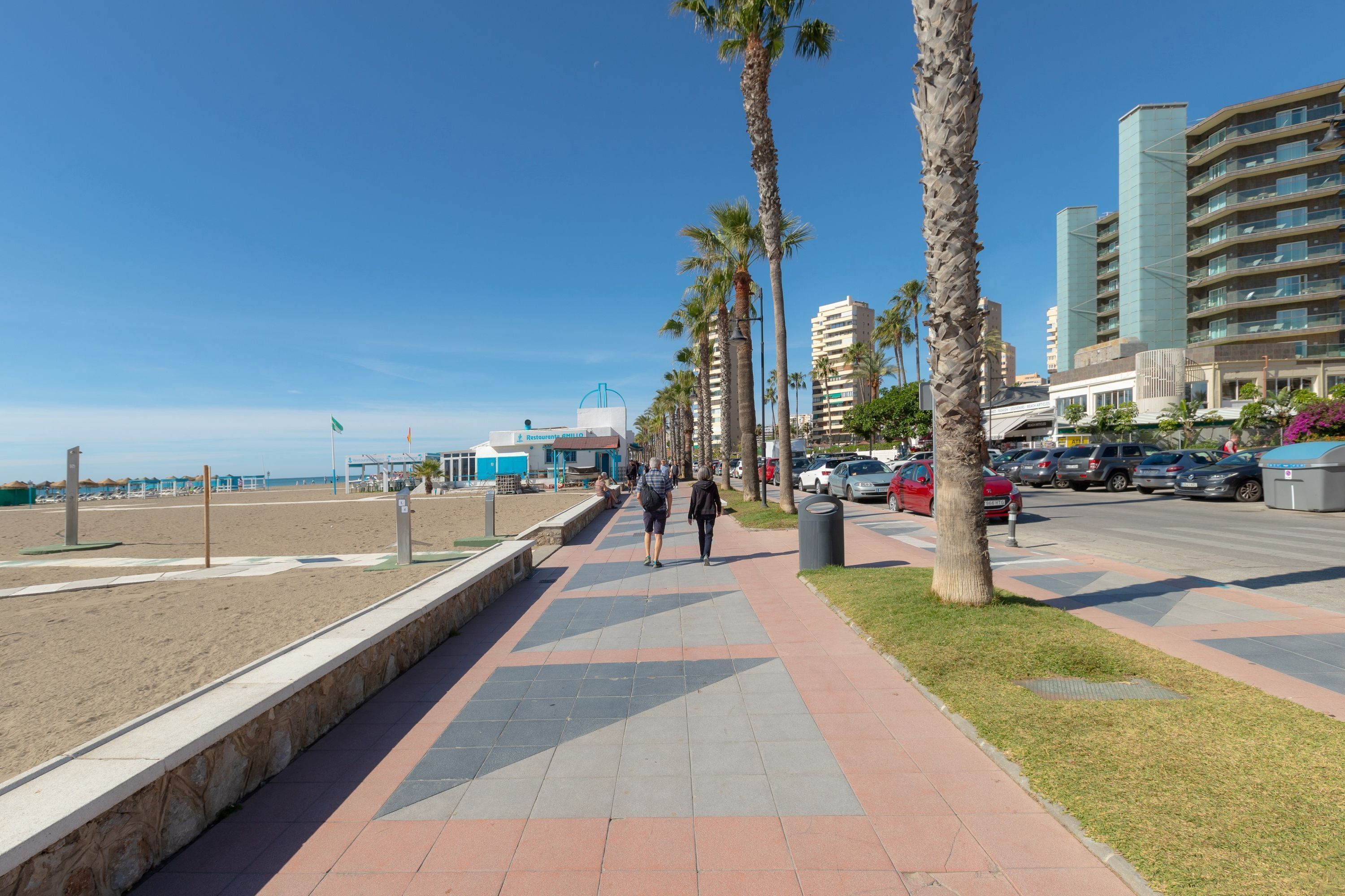 Ein Küstenweg mit Palmen, einem Strand und modernen Gebäuden im Hintergrund.
