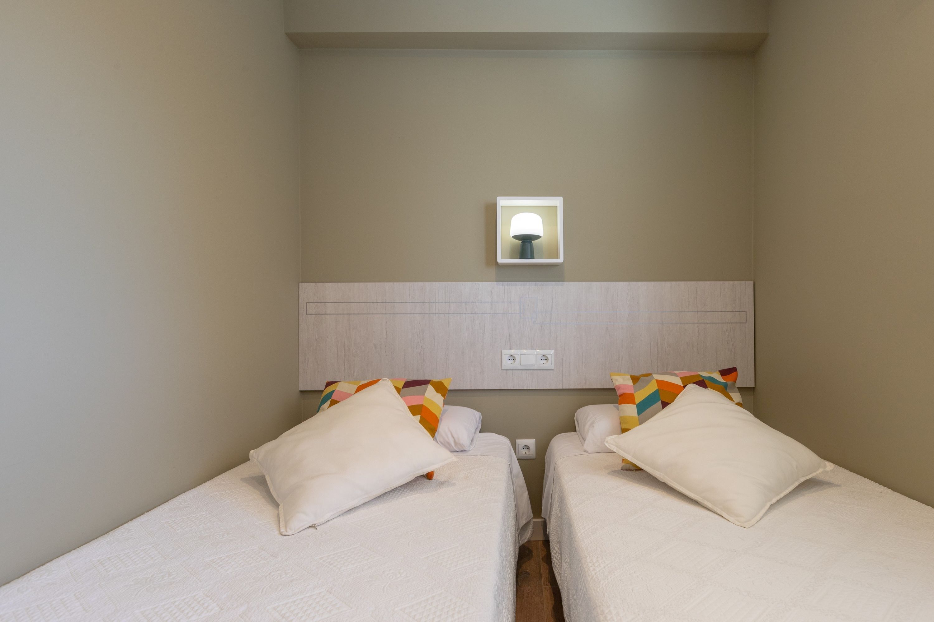 Une chambre d’hôtel avec deux lits simples, une table de chevet, une lampe de chevet, un lampadaire fixé au mur et un mur sobre.