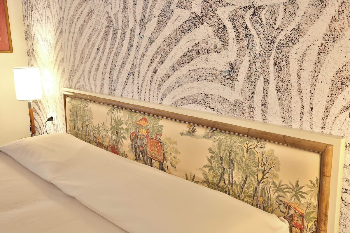 A bed with a headboard featuring a tropical scene, a lamp on the nightstand, and a patterned wall.