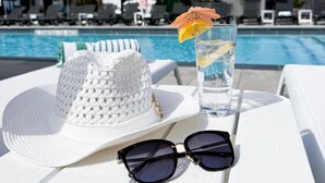 A poolside table with a white hat, sunglasses, and a glass of water with a lemon slice and an umbrella. - Colonial Hotel & Suites - Brenner Premium King (Grand Bend)