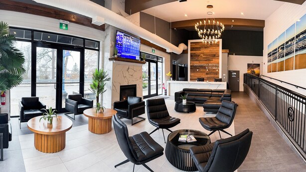 A modern hotel lobby with a fireplace, comfortable seating, a large TV screen, and a view of the outdoors. - Colonial Hotel & Suites - Brenner Premium King (Grand Bend)