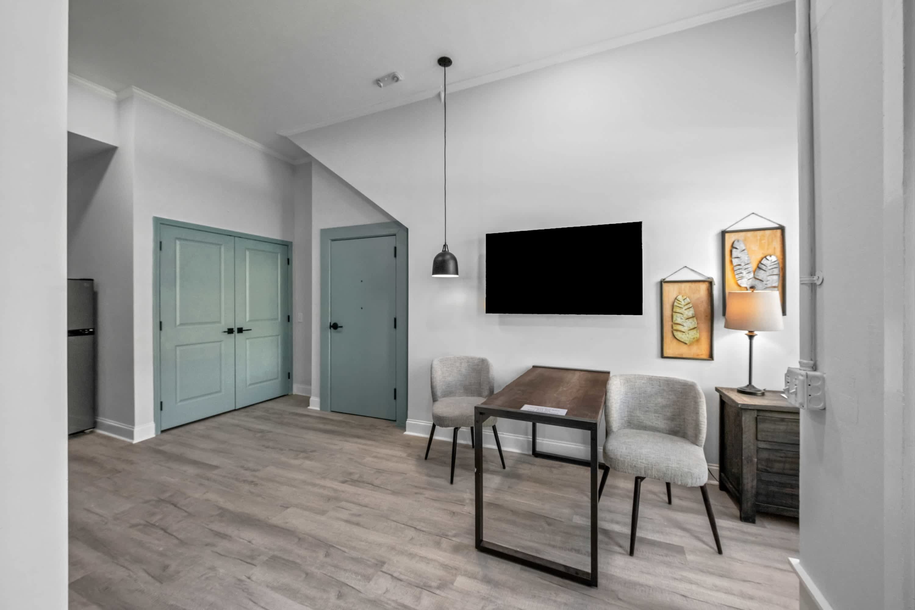 A modern living room with a wooden dining table, two grey chairs, a mounted TV, and a side table with a lamp.