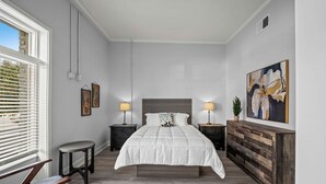 A bedroom with a bed, bedside tables, a nightstand, a chair, a window with blinds, and a painting on the wall.