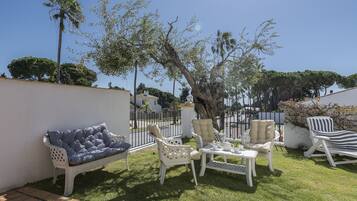 A patio with white furniture, including a sofa, chairs, and a table, set against a backdrop of trees and a clear sky.