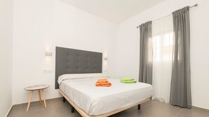 A neatly made bed with a grey headboard, a small round table with a patterned top, and grey curtains on a window. - Miamar 1 in Hozanejos (Chiclana de la Frontera)