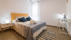 Un dormitorio con una cama, una mesita de noche con lámpara, un armario blanco pequeño y una ventana con cortinas.