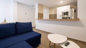Una sala de estar moderna con un sofá azul, una mesa blanca y una zona de cocina con microondas y refrigerador.