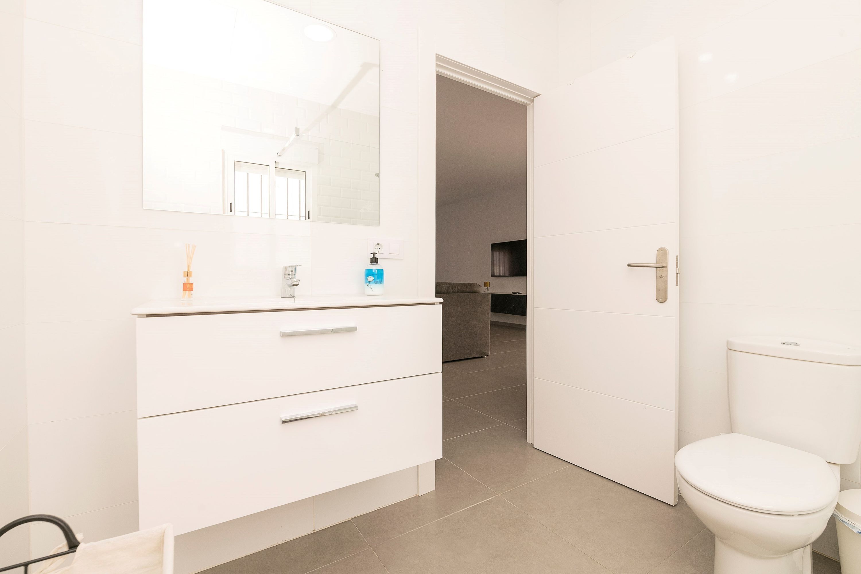 Une salle de bain moderne avec un meuble-lavabo blanc, un grand miroir, des toilettes et un espace douche visible à travers une porte ouverte.