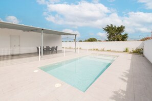 Una piscina con solárium de baldosas blancas, una zona de estar cubierta y un edificio blanco con una puerta.