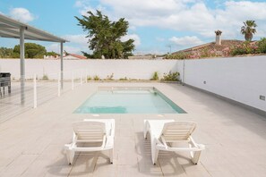Una piscina con dos sillas reclinables blancas frente a ella, rodeada por un muro blanco y una zona techada.