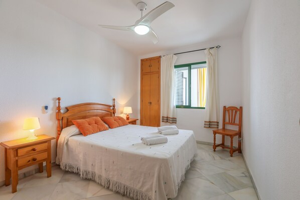 Une chambre à coucher comprenant un lit, des tables de chevet, une chaise en bois, un ventilateur de plafond et une fenêtre avec des rideaux.
