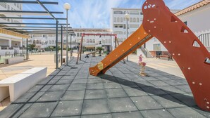 En udendørs legeplads med en stor orange rutchebane og gynger, omgivet af bygninger og en klar himmel.