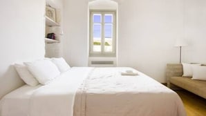 A bedroom with a large bed, a sofa, a window with a view, and a shelf with items.