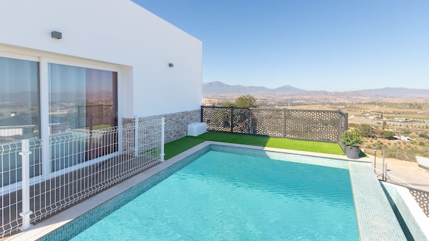Un espace piscine moderne offrant une vue dégagée sur les montagnes lointaines, un bâtiment blanc doté de grandes fenêtres et une petite pelouse.