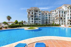 A swimming pool with lounge chairs, surrounded by multi-story buildings with balconies and a clear blue sky. - Marina De La Duquesa 805 (Manilva)