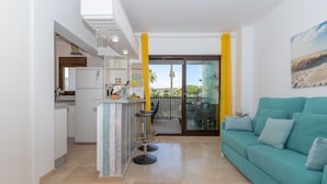 A modern kitchen with a refrigerator, a bar stool, and a dining area with a table and chairs. There is a teal sofa and a painting on the wall. - Marina De La Duquesa 805 (Manilva)