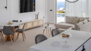 Un soggiorno moderno con un divano, un'area pranzo con tavolo e sedie, una TV a parete e vista sull'esterno attraverso ampie finestre.