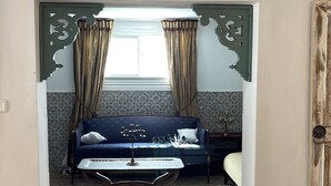 Ein Zimmer mit einem blauen Sofa, zwei Poufs, gemusterten Teppichen und einem Fenster mit Vorhängen.
