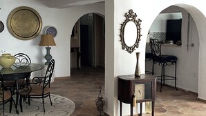 Ein großzügiger Wohnbereich mit einem runden Tisch, Stühlen, einer Couch und einem Couchtisch. An der Wand befinden sich dekorative Teller und eine Lampe. Der Boden ist mit Fliesen ausgelegt, und es gibt zwei Bogenportale, die in andere Räume führen.