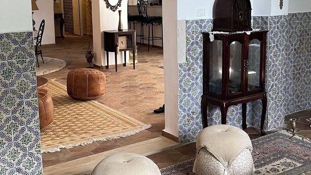 Ein geräumiger Wohnbereich mit einer großen Couch, Hocker und einem gemusterten Teppich. Es gibt einen Holzschrank mit Glastüren und einen dekorativen Spiegel an der Wand.