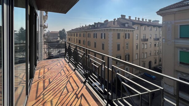 A balcony with a tiled floor and a metal railing overlooking a cityscape with buildings and a clear sky. - [Exclusive 120 SQM] in the heart of Bergamo (Bergamo)