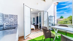 En moderne udendørs terrasse med spisebord og stole, et lille tekøkken med vask, og udsigt til blå himmel og grønt.