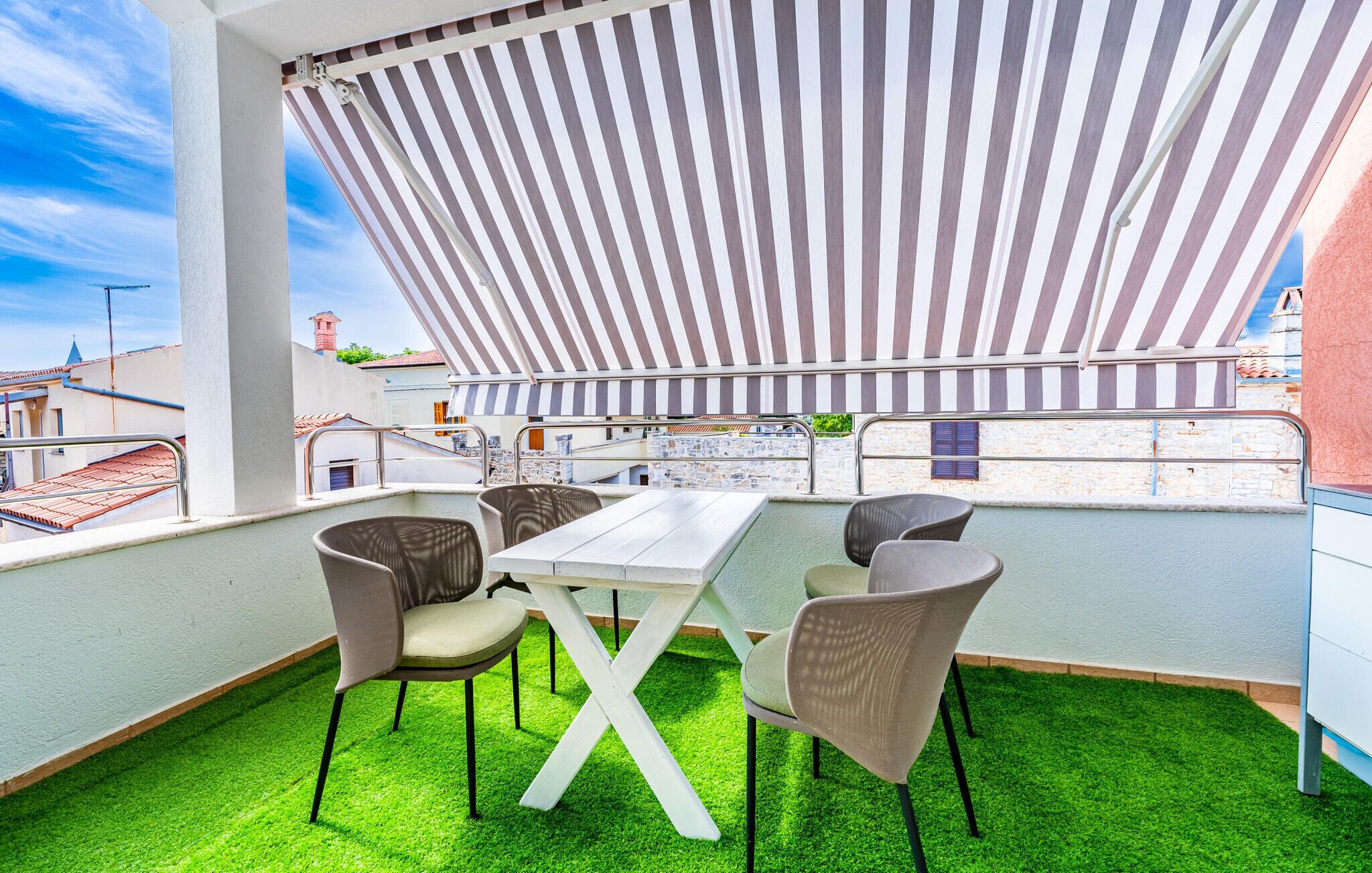 Una terraza al aire libre con una mesa blanca y cuatro sillas, rodeada por un toldo a rayas y césped artificial verde.