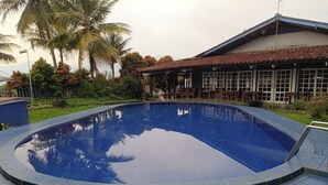 A swimming pool with a tiled edge, surrounded by a grassy area and palm trees. - Capital O 94094 New Lembah nyiur (Cisarua)