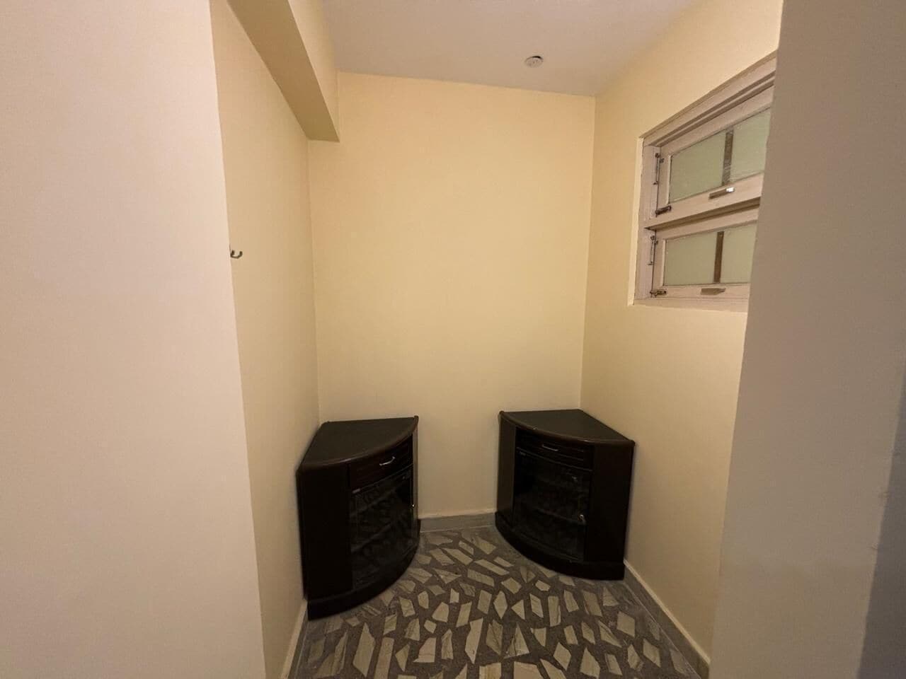 A narrow hallway with two dark-colored cabinets, a patterned floor, and a window with blinds.