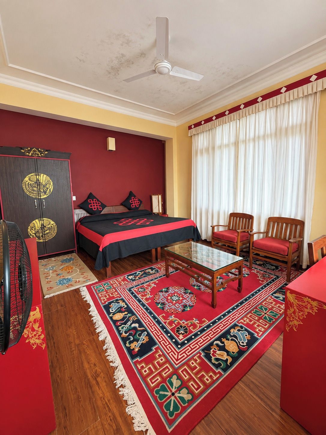 Habitación de hotel con cama, escritorio, dos sillas, ventilador de techo, armario y ventana con cortinas.