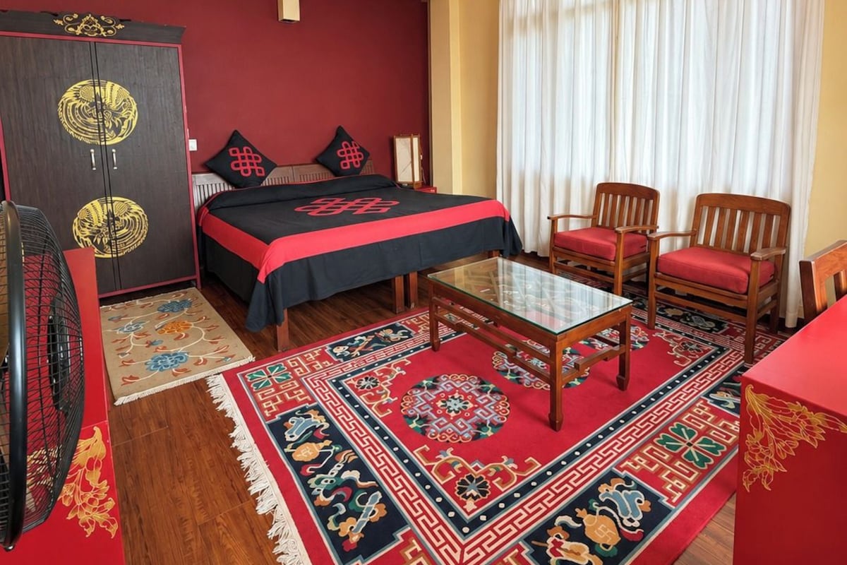 A hotel room with a bed, a desk, two chairs, a ceiling fan, a wardrobe, and a window with curtains.