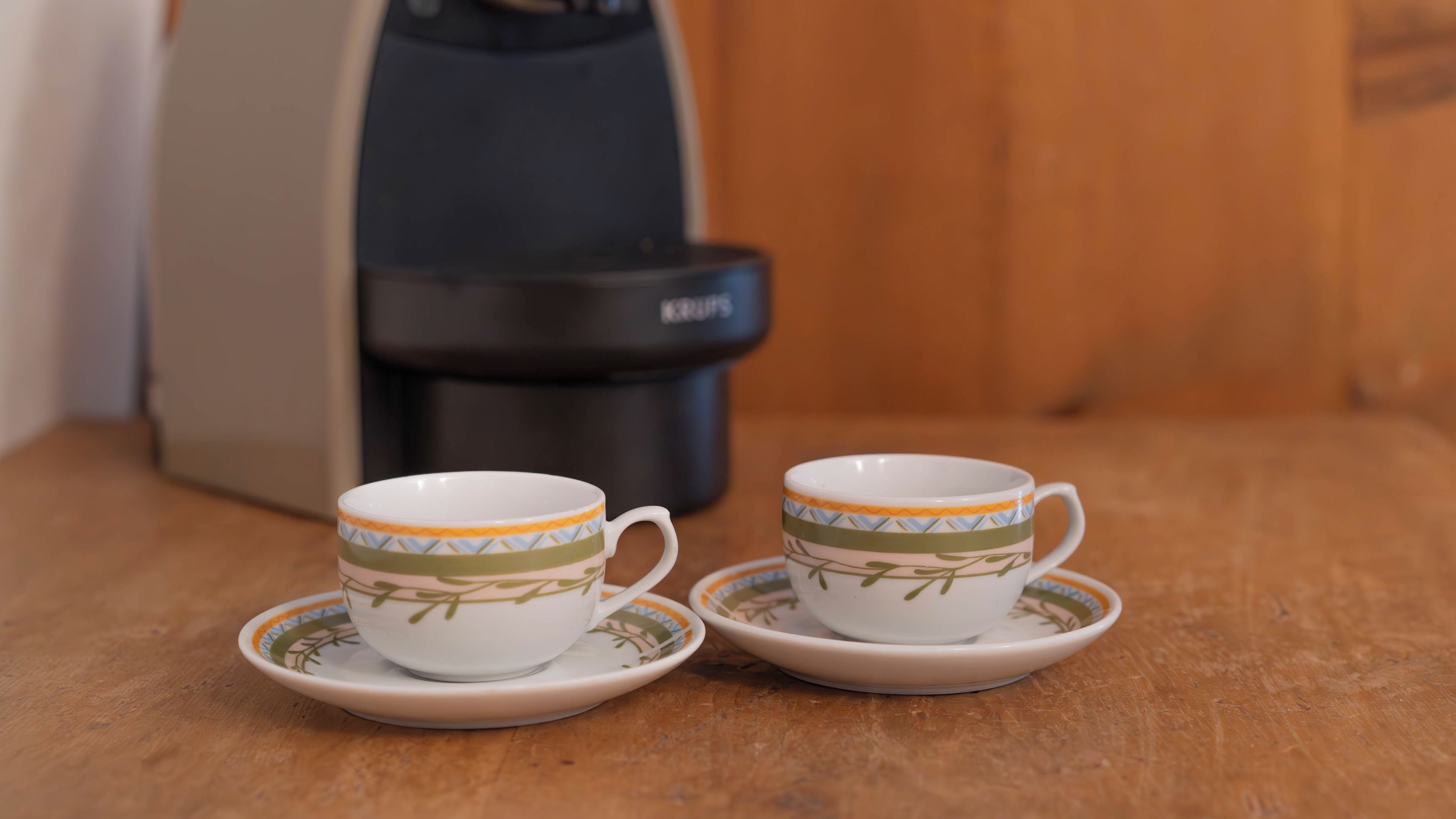 Two white cups with green leaf patterns on saucers, placed on a wooden table.