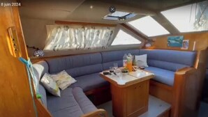A yacht interior with a blue cushioned seating area, a small wooden table with drinks, and a window with curtains. - Boat rental (Marseille)