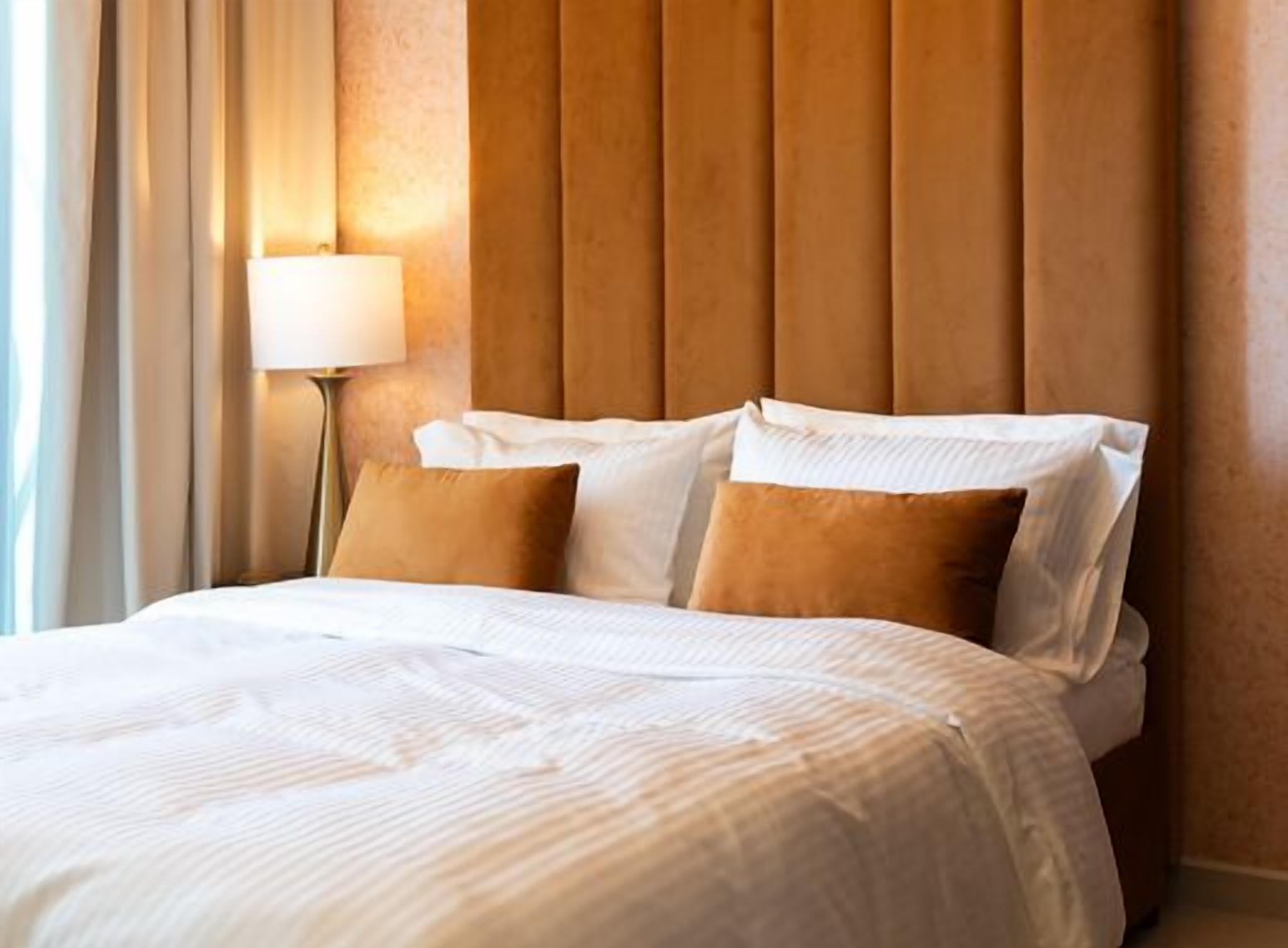 A neatly made bed with white linens and two brown pillows, a wooden headboard, and a bedside lamp.