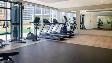 Une salle de sport moderne équipée de tapis de course, d’une machine de musculation et d’un banc. De grandes fenêtres laissent entrer la lumière naturelle et offrent une vue sur la ville.