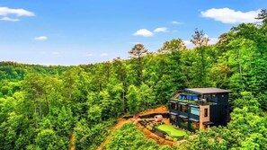 A modern two-story house surrounded by dense forest, with a visible deck and outdoor seating area. - Rooftop Heated Pool - Fire Pit - Pool table - LUX (Gatlinburg)